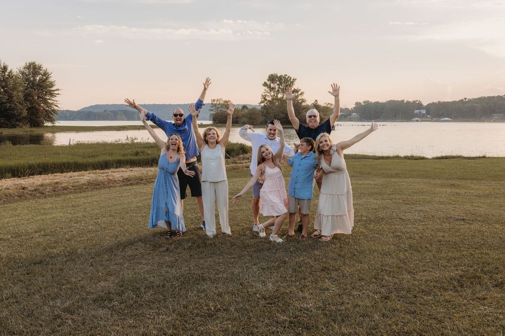Group of joyful people celebrating outdoors by a lake at sunset, arms raised in excitement on green grass.