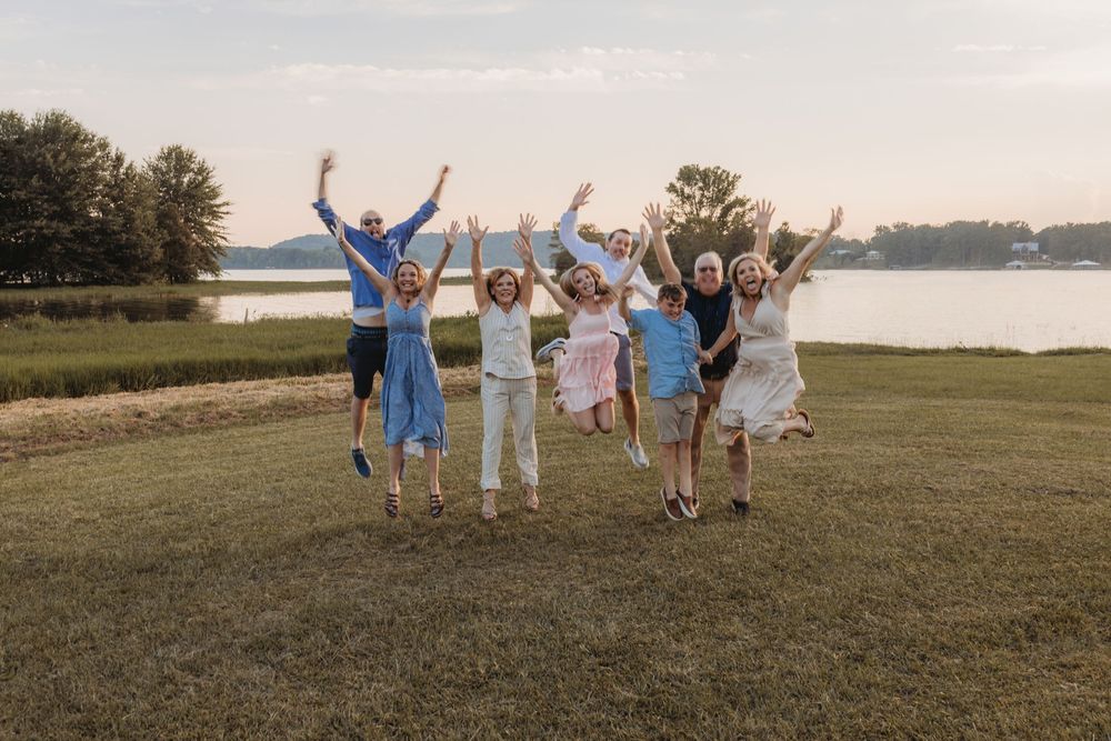 A joyful group of seven people jumping with arms raised on a green lawn by a lake at sunset.