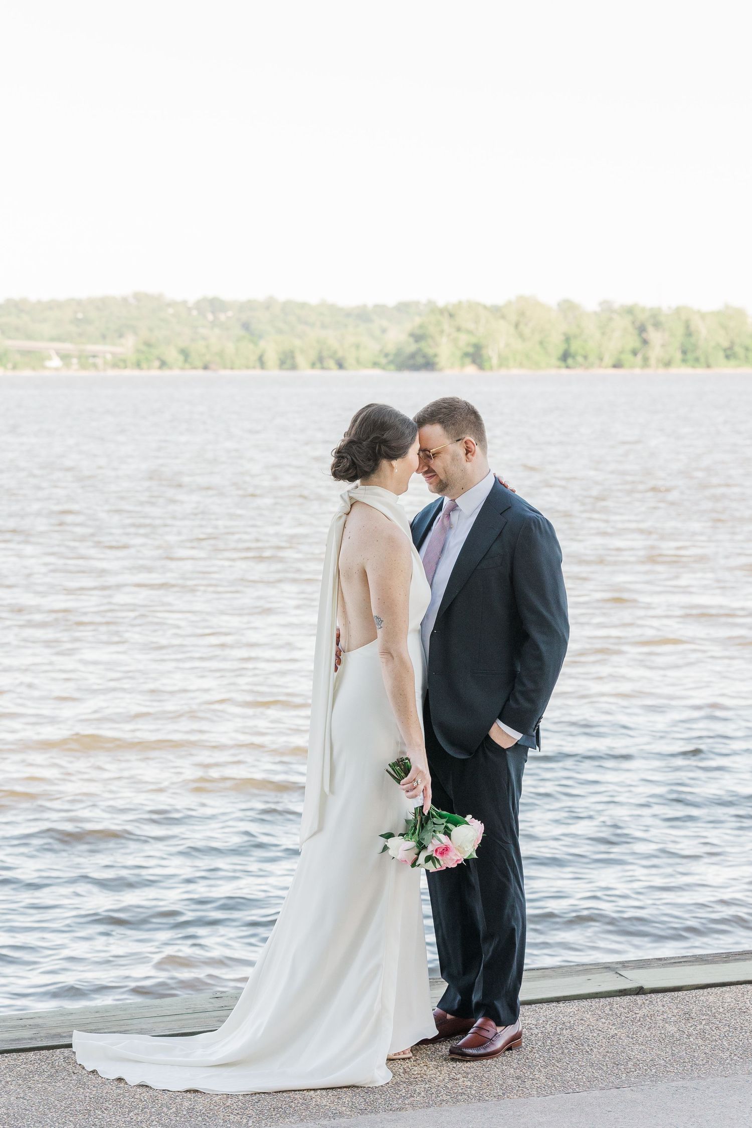 Bride and groom share a romantic moment by the waterfront, bride in elegant backless gown holding pink bouquet.