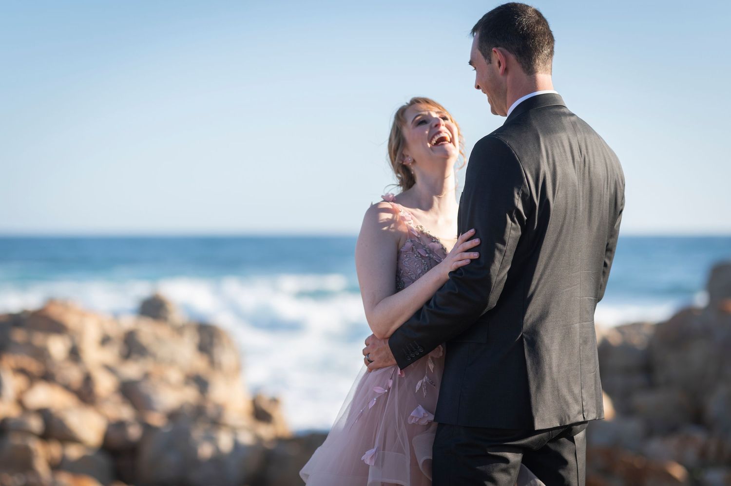 Couple embracing on rocky ocean shore, woman in pink gown smiling at man in gray suit with waves behind them.