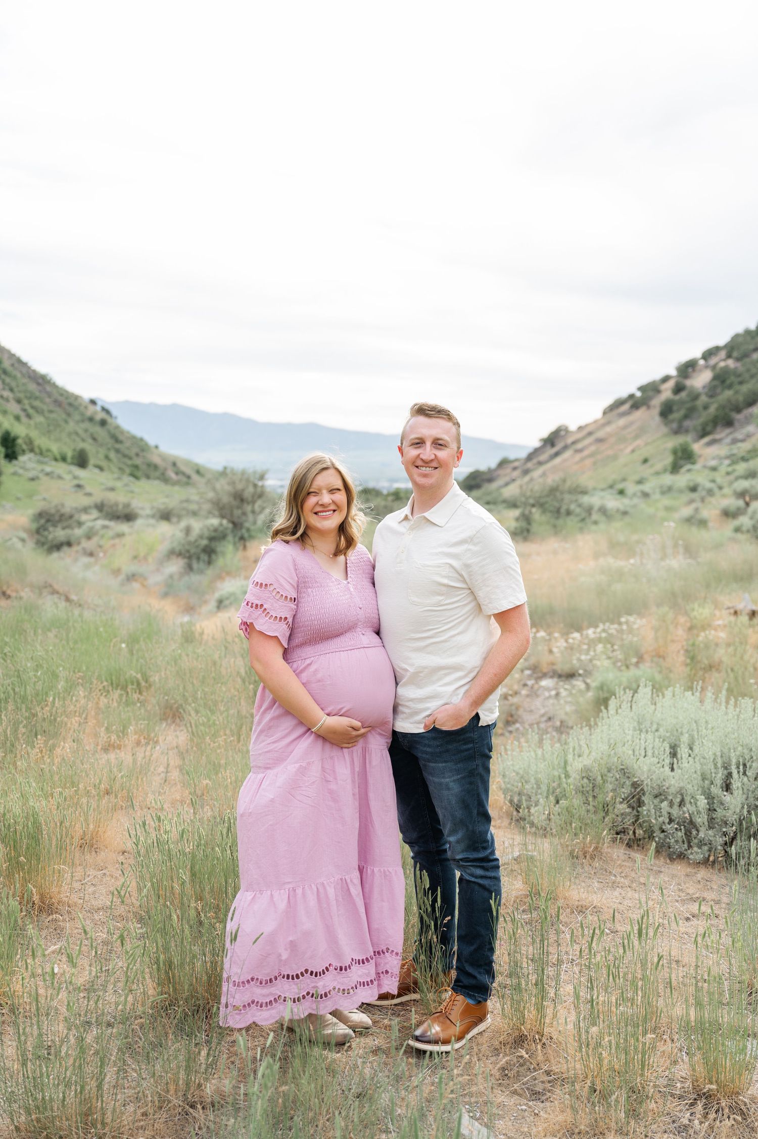A couple poses in a scenic mountain valley with sage brush, the woman wearing a flowing pink maxi dress.