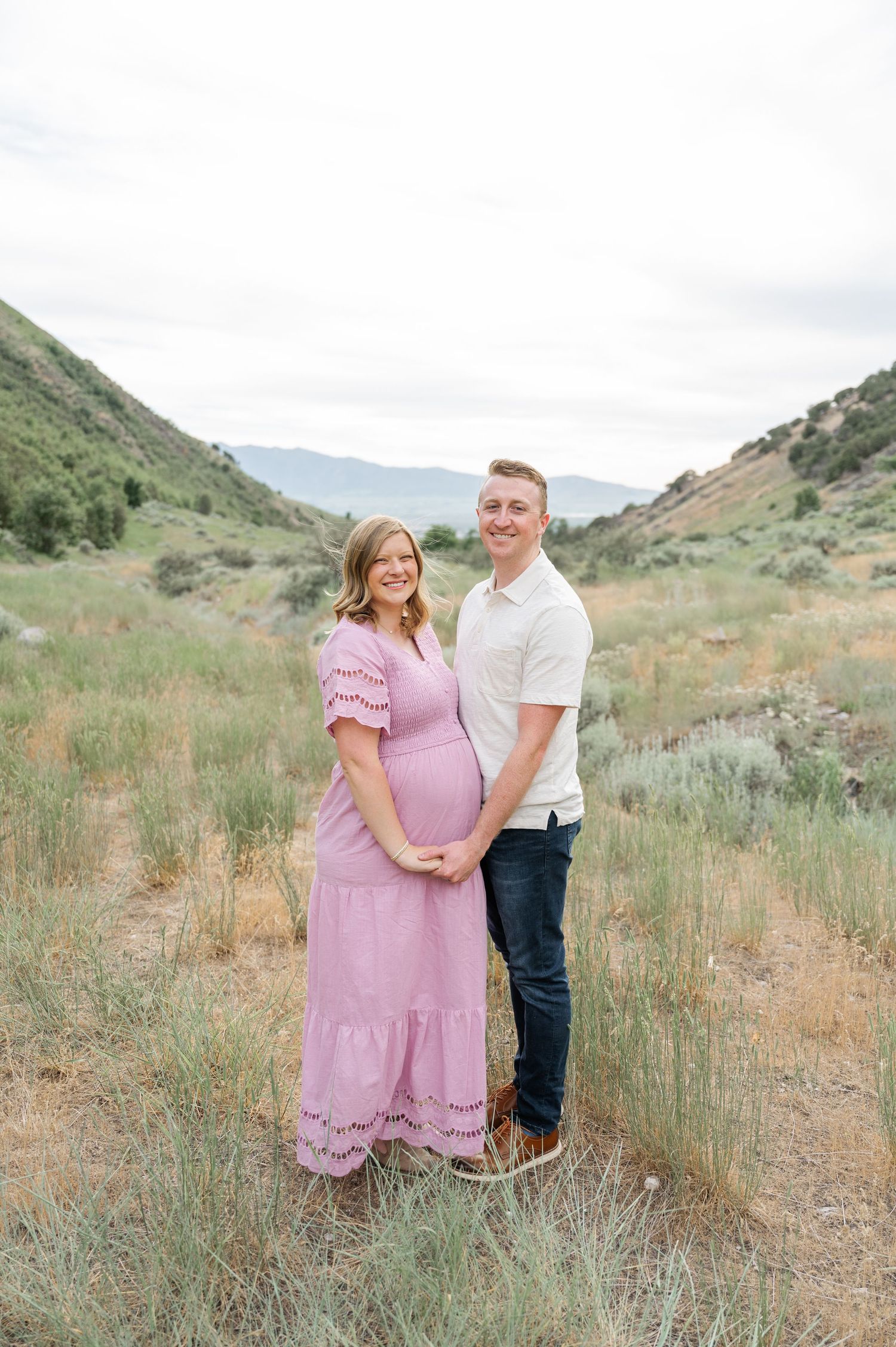 A couple poses together in a scenic mountain valley setting with green hills, the woman wearing a pink maxi dress.