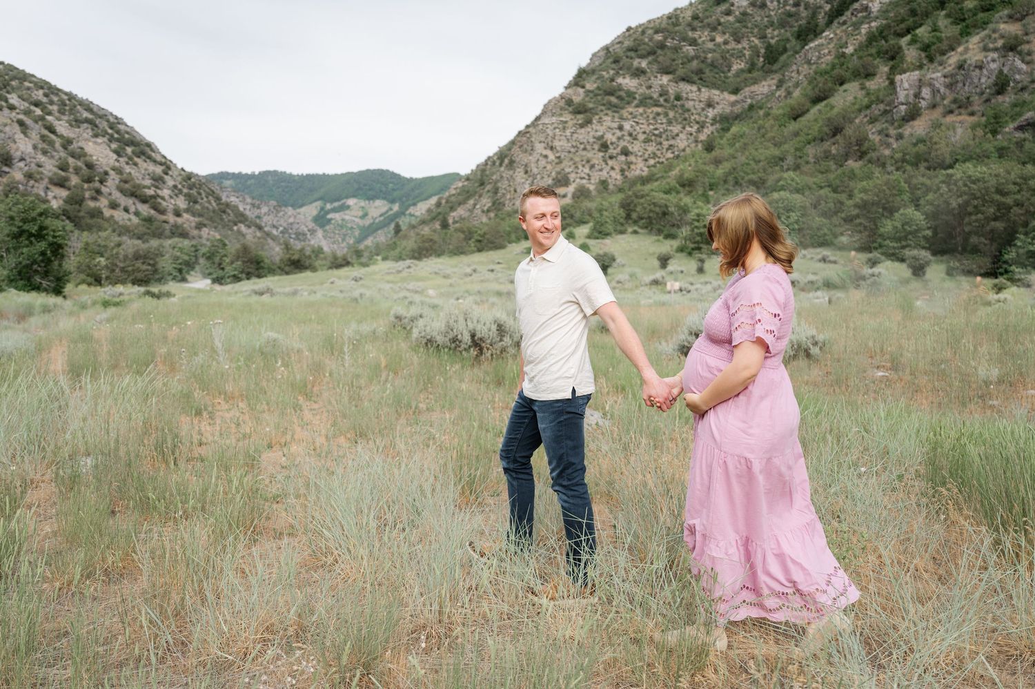A couple poses in a grassy meadow surrounded by mountains during a maternity photo shoot on a sunny day.