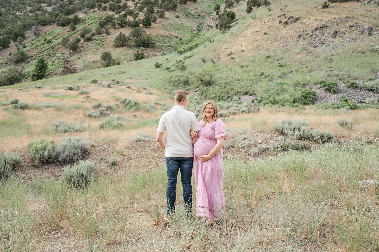 A couple stands together on a hillside covered in sage brush and native grasses during golden hour.