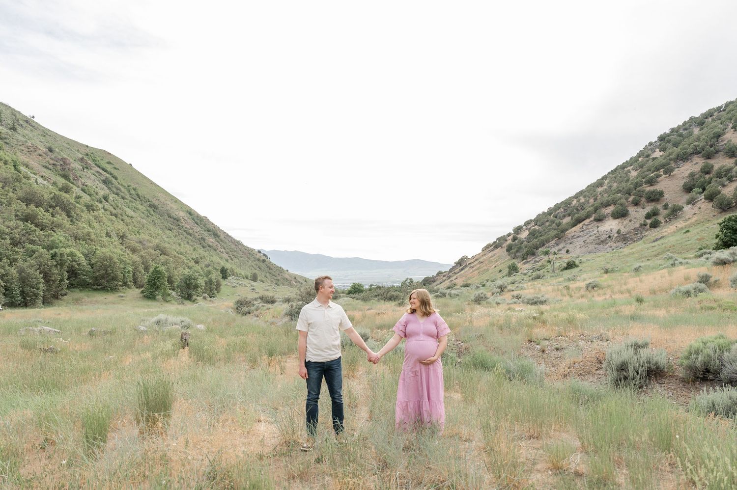 Two people stand together in a scenic mountain valley surrounded by rolling hills and sagebrush.
