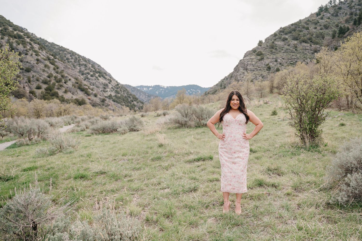 A person in a light colored dress stands in a grassy mountain valley surrounded by rugged terrain and desert brush.