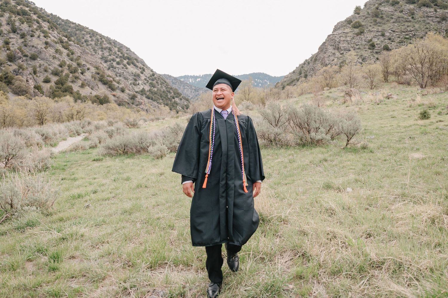 A graduate in traditional black cap and gown stands in a grassy mountain valley on their graduation day.