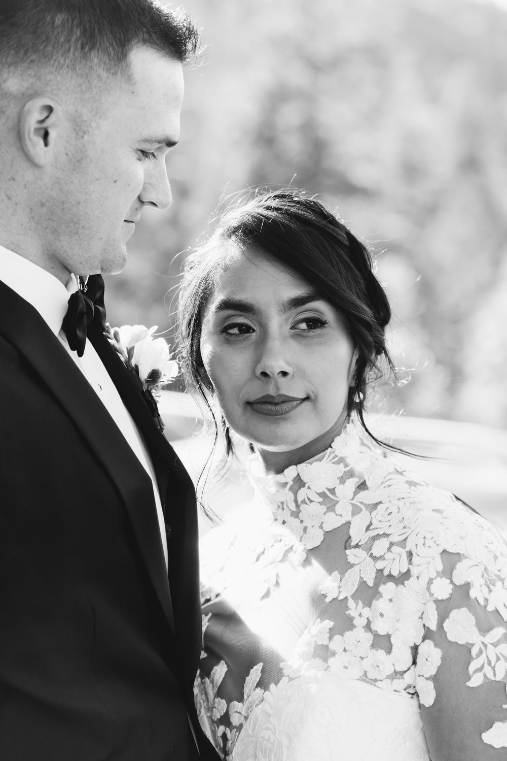 Black and white wedding portrait of bride in lace gown gazing at camera beside groom in tuxedo with bow tie.