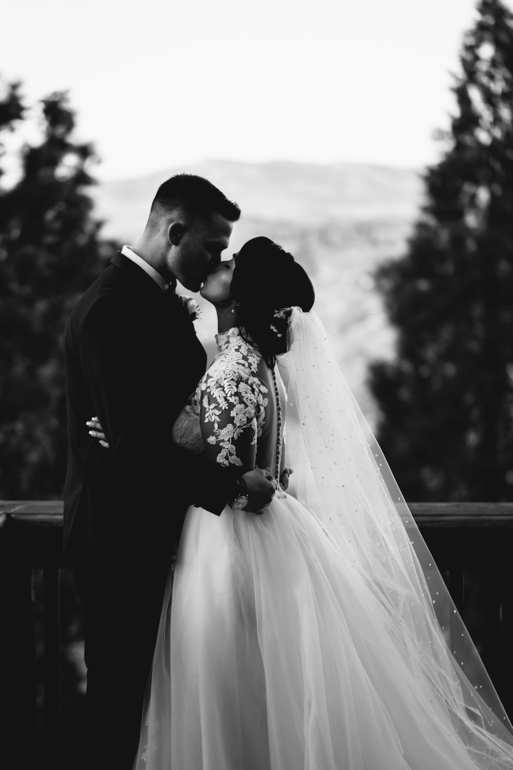Black and white wedding photo of bride in lace gown and veil embracing groom in suit outdoors.