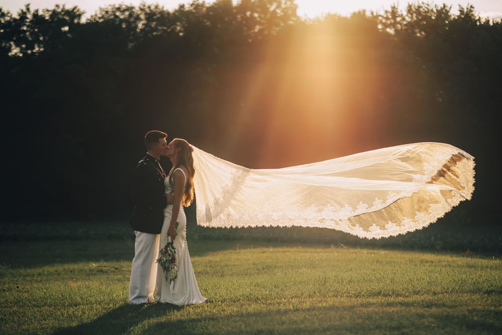 Couple sharing a romantic kiss at sunset, bride's long veil flowing in the wind on a lush green lawn.