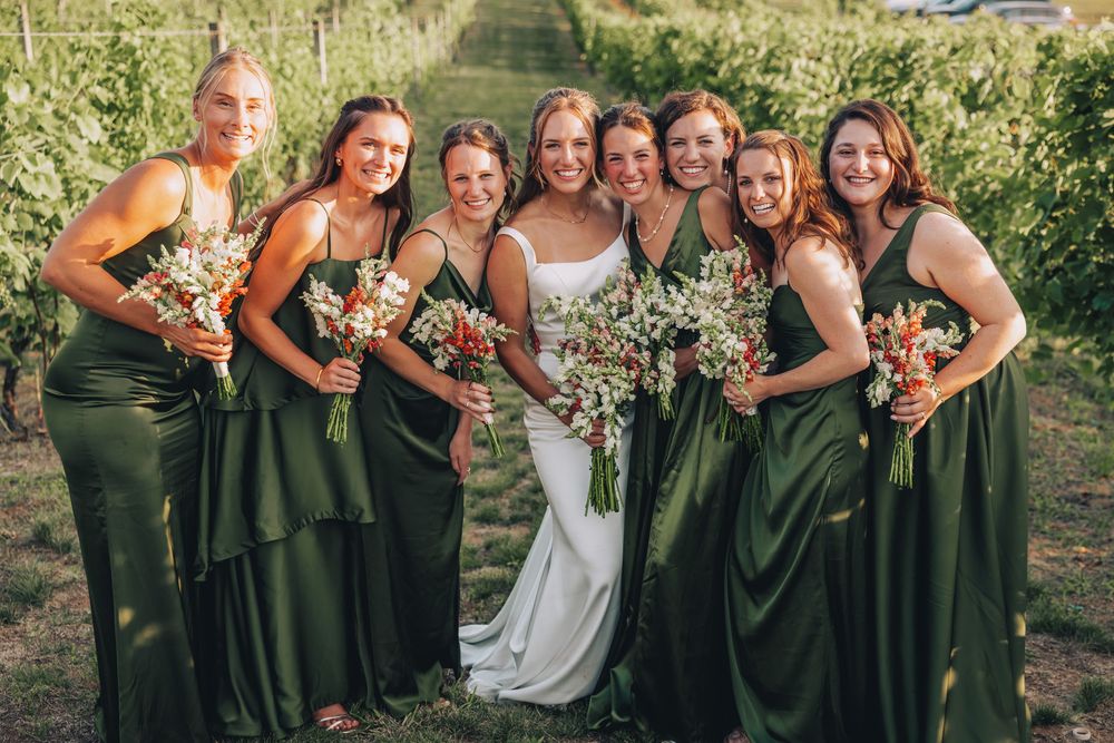Bride in white gown with seven bridesmaids in olive green dresses holding bouquets in a vineyard.