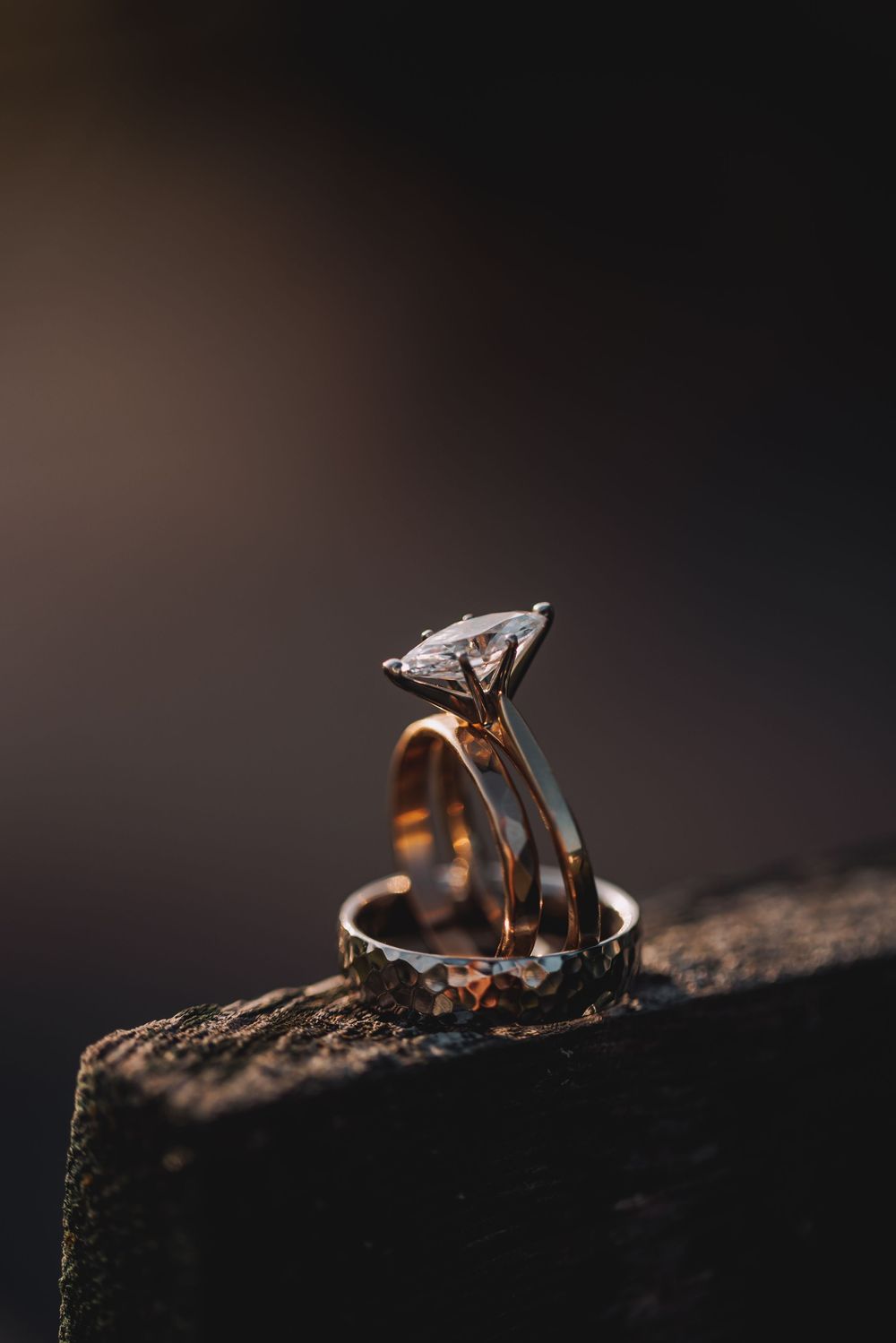 Gold engagement ring with large diamond solitaire and wedding bands stacked on dark rustic surface.