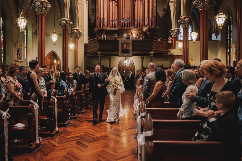 Bride walking down the aisle of an ornate church with wooden pews and pipe organ during a wedding ceremony.