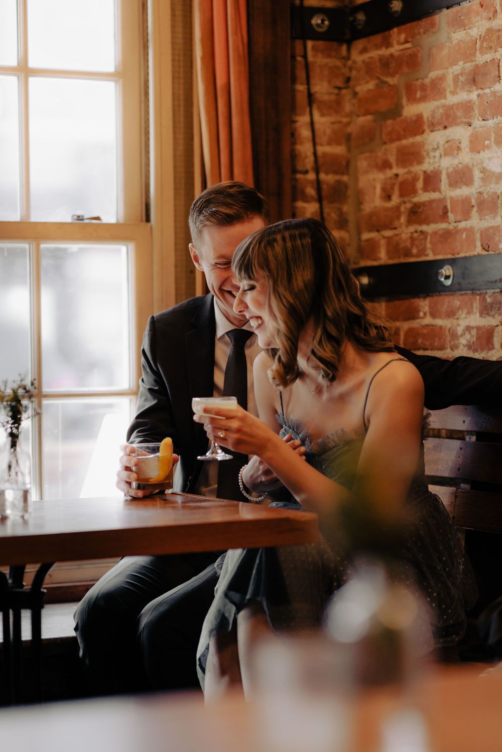 A couple laughs together over cocktails at a cozy brick-walled restaurant, dressed in elegant attire.