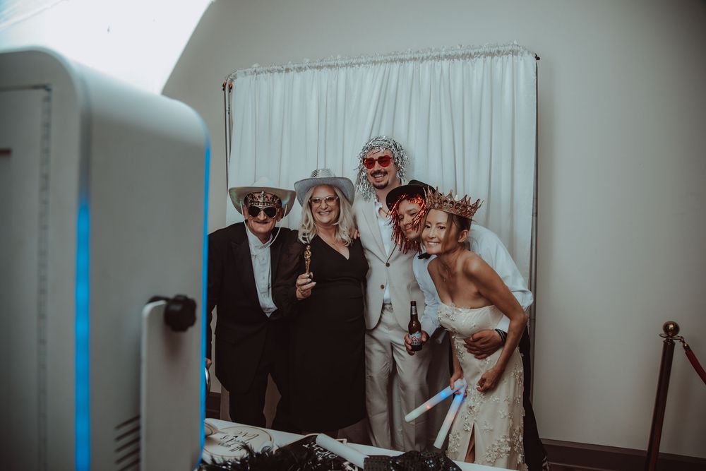 Wedding guests pose in fun costumes and props at a photo booth with white curtain backdrop.