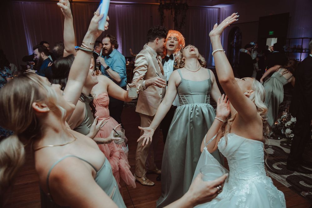 Bride and wedding guests dance joyfully on the reception dance floor with hands raised.