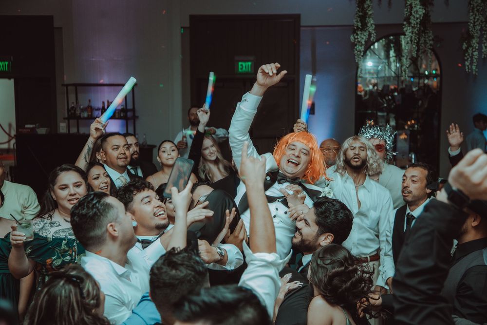Energetic wedding reception dance floor crowd celebrating with glow sticks and colorful wigs.
