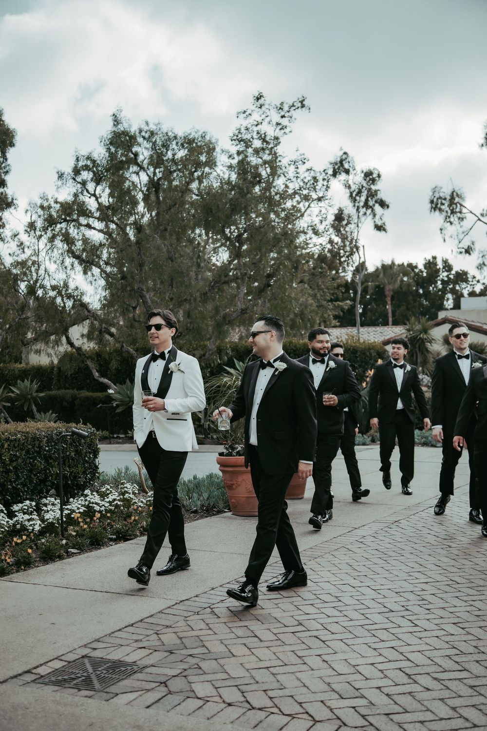 Groom in white tuxedo jacket leads groomsmen in black tuxedos along an outdoor wedding venue path.