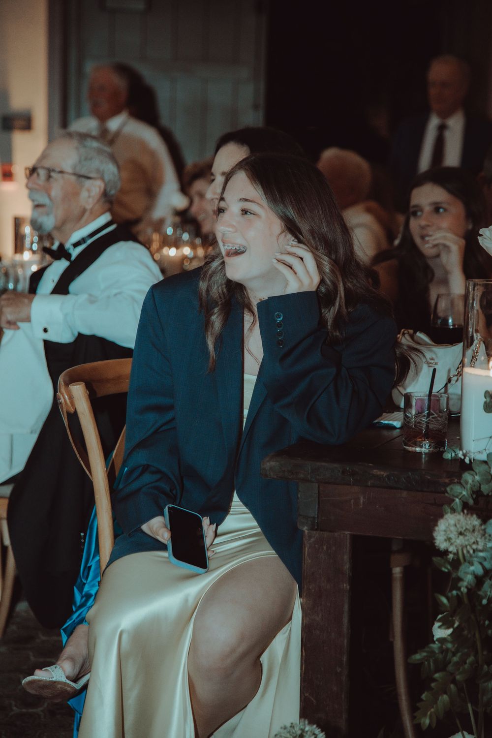 Wedding guest in a navy blazer and satin skirt smiles while watching the reception festivities at a candlelit venue.