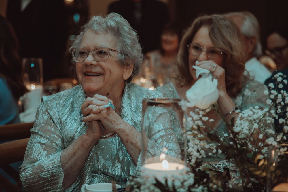 Elderly woman with clasped hands smiles joyfully at a wedding reception with candles and floral centerpieces.
