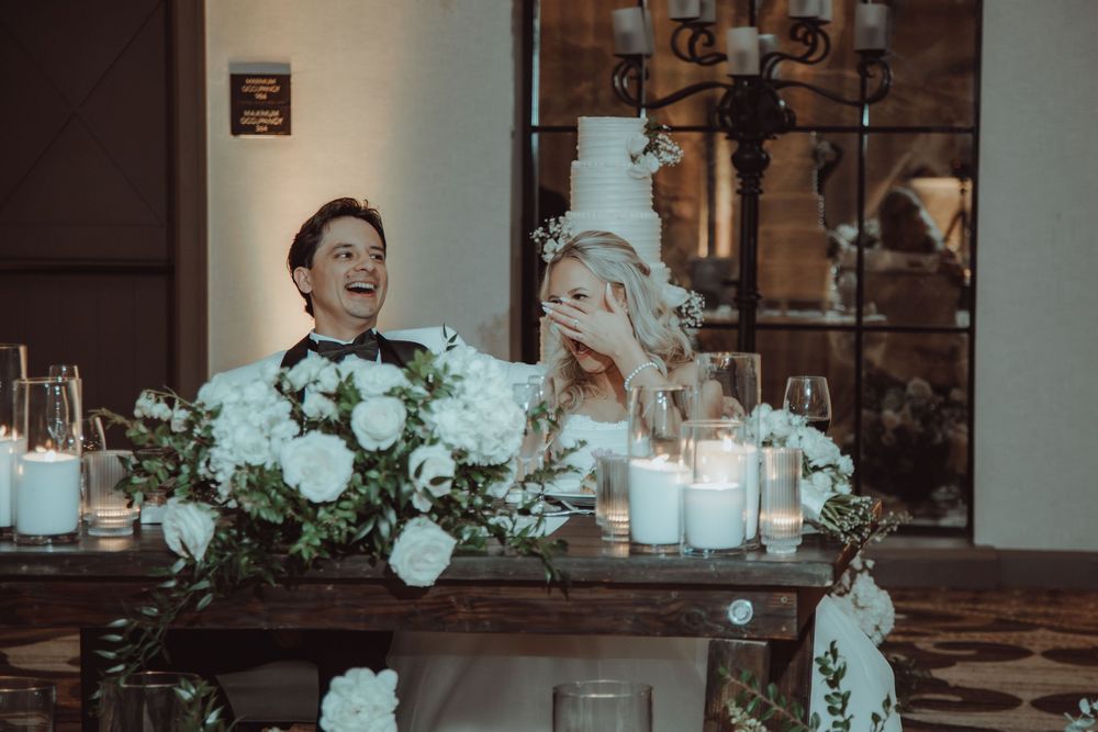 Bride and groom laugh together at the sweetheart table surrounded by white floral arrangements and candles.