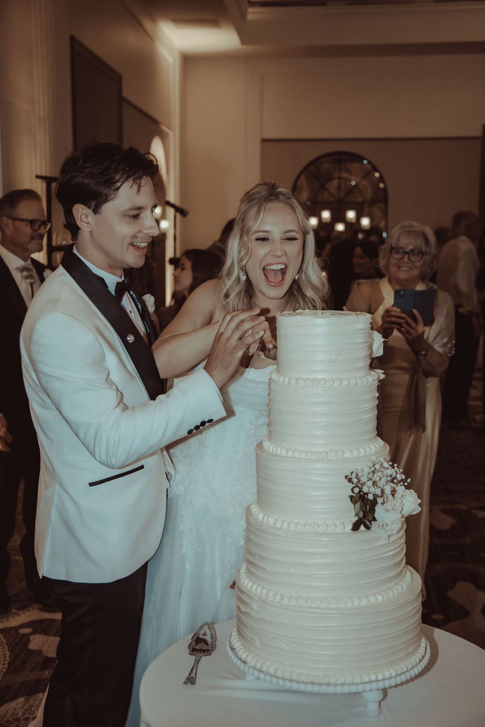 Bride and groom joyfully cut their tall four-tier white wedding cake at their elegant reception.