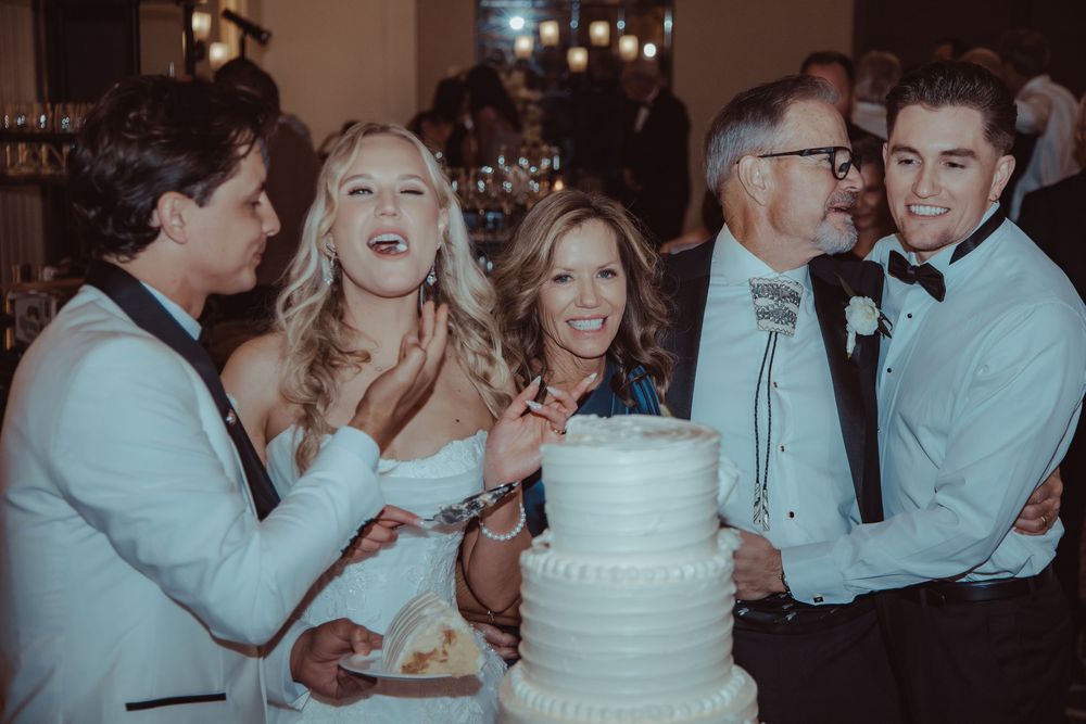 Bride, groom, and family members gather around the white wedding cake smiling during the reception celebration.