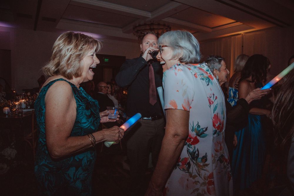 Two women laugh and dance together holding glow sticks at a fun and lively wedding reception.