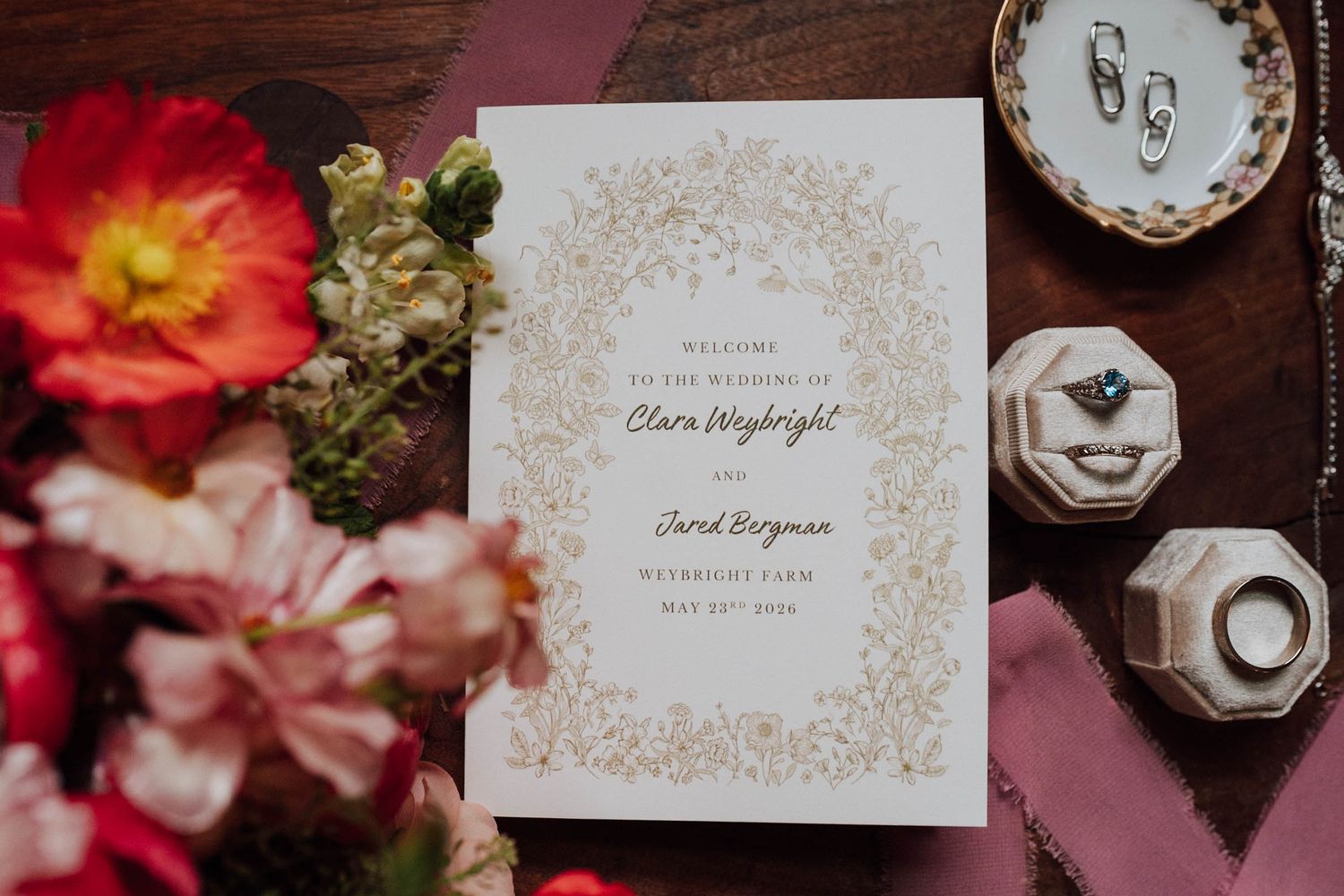 Elegant wedding program with gold floral border beside rings, earrings, and fresh flowers on a wooden surface.