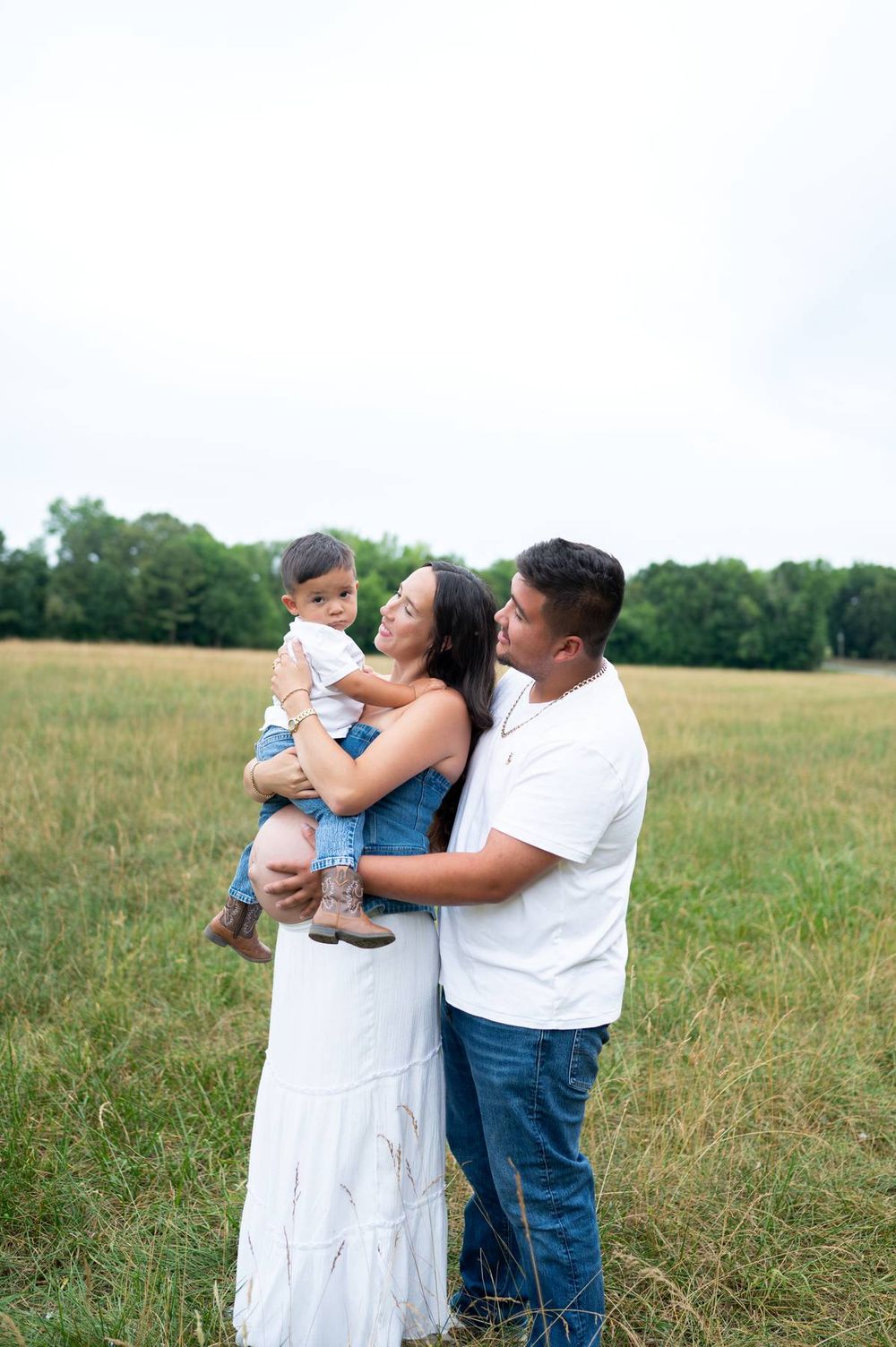 Family maternity portrait in open field, mother holding toddler while father embraces her in North Carolina