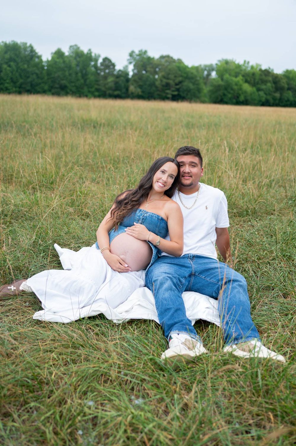 Smiling pregnant couple sitting together on white blanket in golden grass field in North Carolina