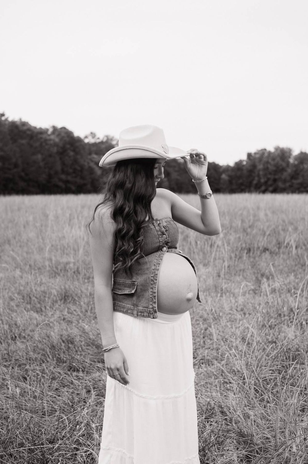 Black and white maternity photo of pregnant woman in cowboy hat and denim vest in field in North Carolina
