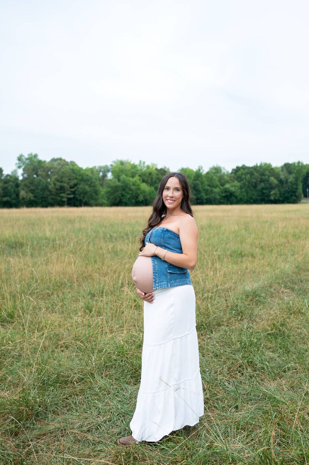 Pregnant woman in denim top and white maxi skirt cradling baby bump in a green field in North Carolina
