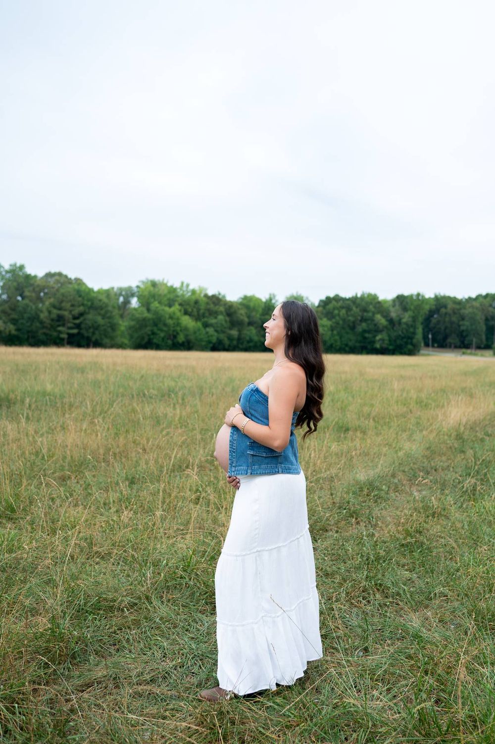 Side profile of pregnant woman in denim tube top and white skirt standing in an open field in North Carolina