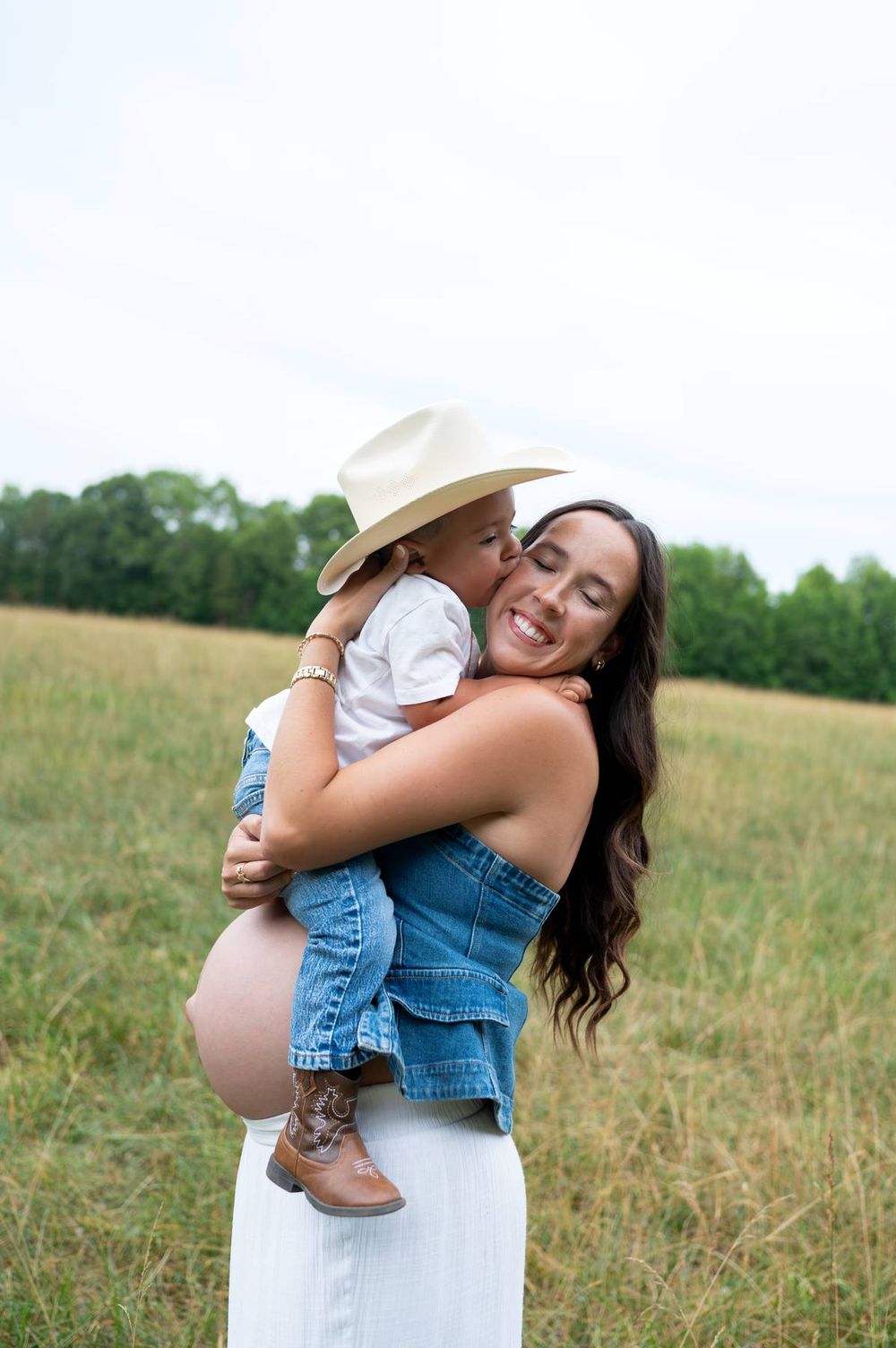 Smiling pregnant woman holding young child in cowboy hat and boots in a grassy field Lexington NC