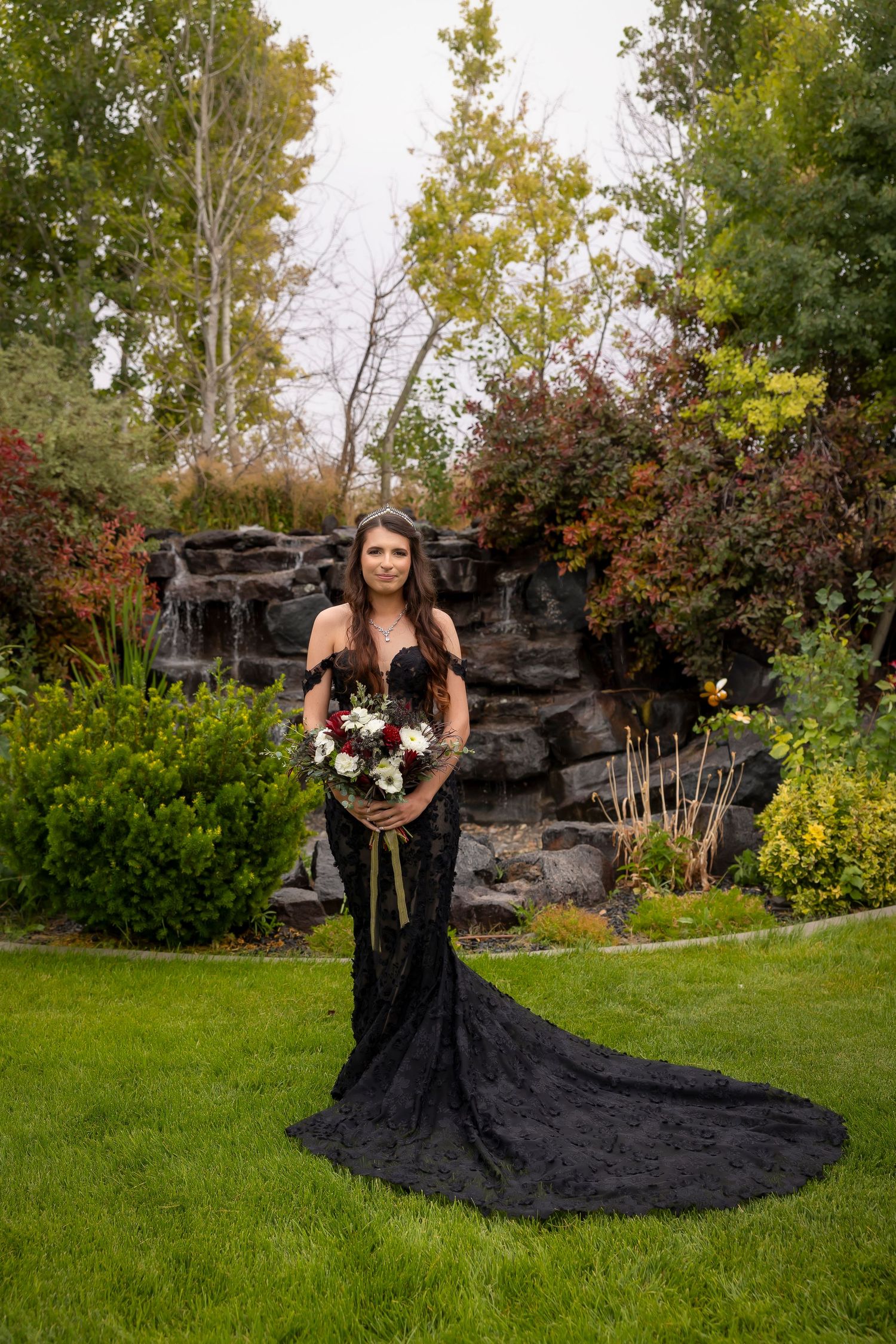 bride wearing black fitted lace gown standing with bouquet in front of waterfall Castle Gardens Kuna Idaho ©Milestones Studios LLC