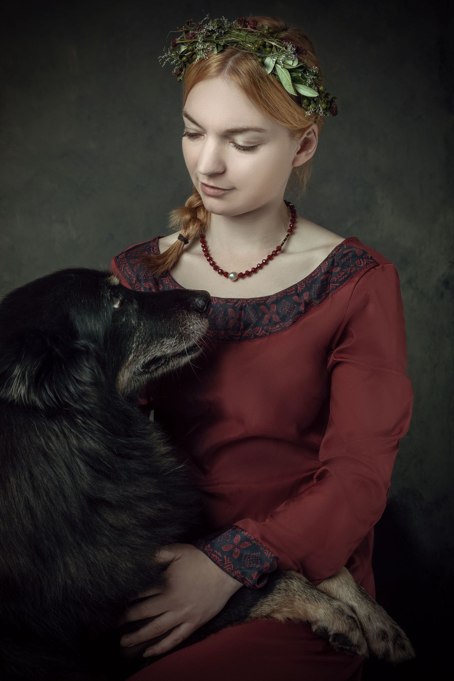 Young woman in red medieval dress with floral crown holds a black dog in a dark, moody portrait setting.