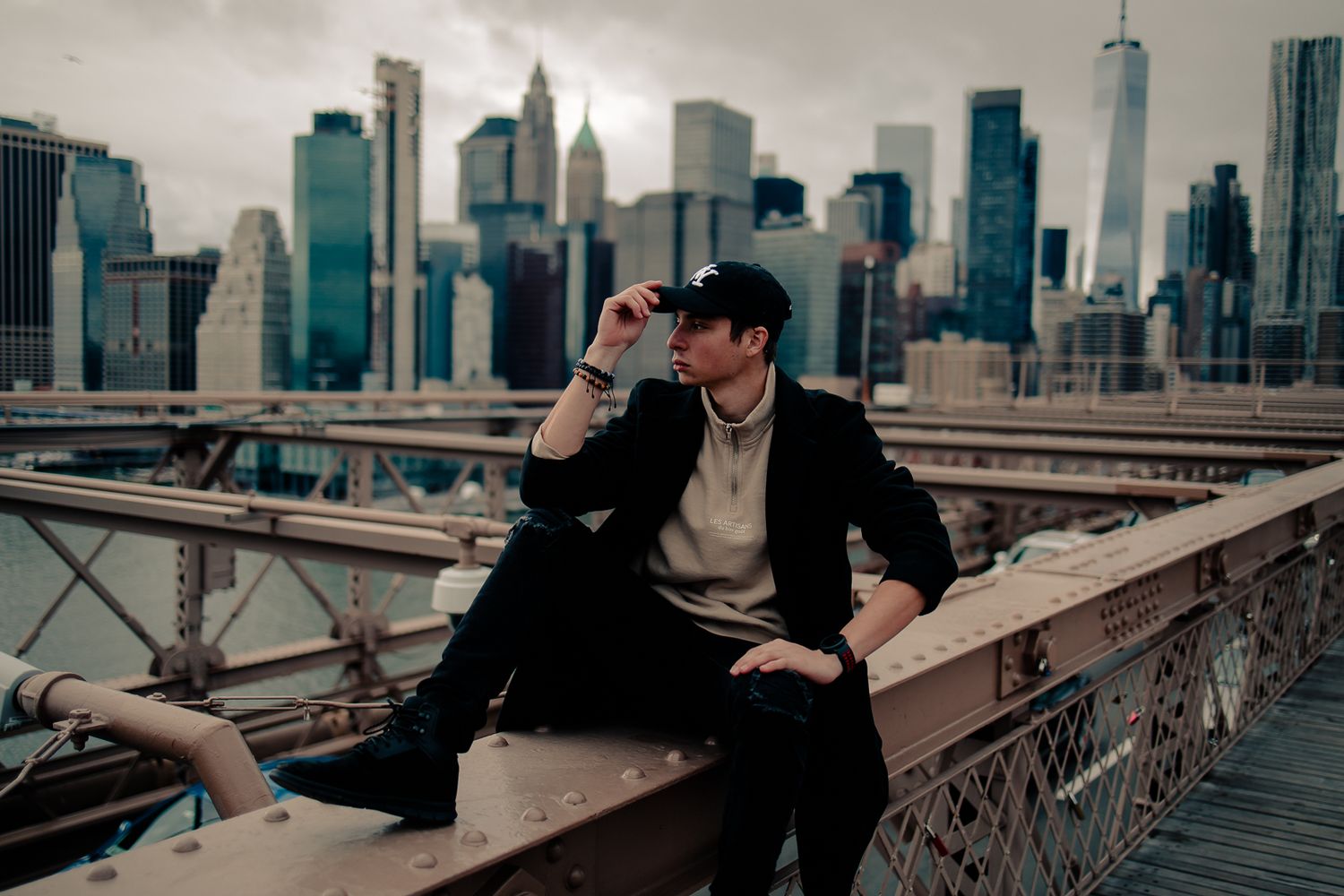 Man in a black coat sitting on the Brooklyn Bridge railing with the Manhattan skyline, New York City | Mannyportraits