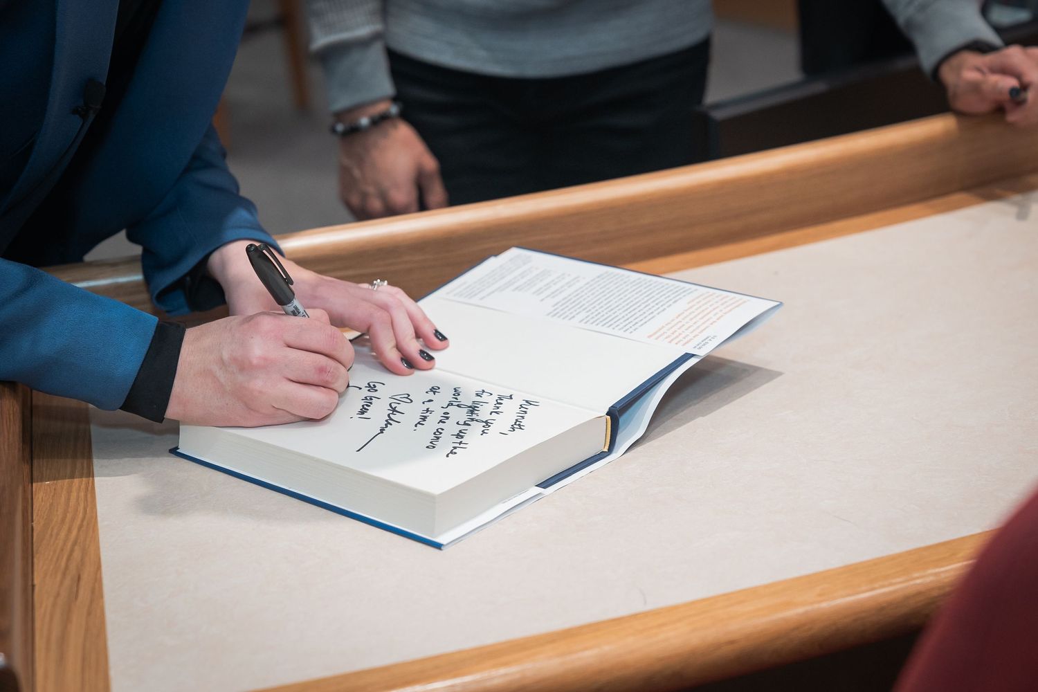Person signing a book with a pen at a wooden table during a book release event.
