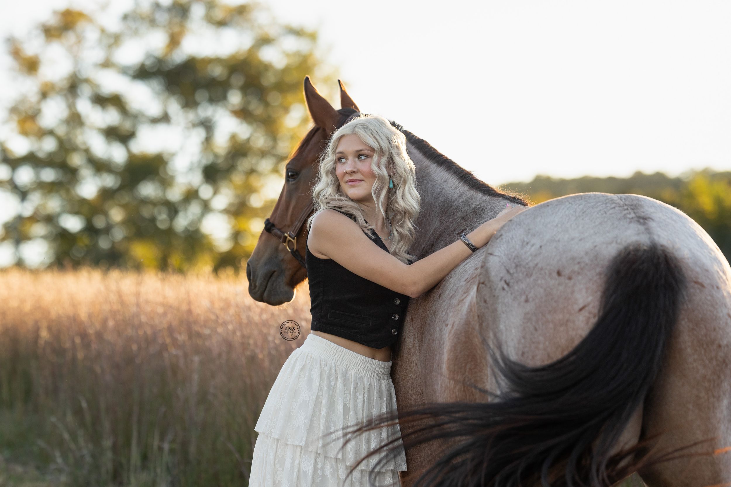 Blonde woman in black top and white skirt embracing a gray horse in a golden field at sunset.