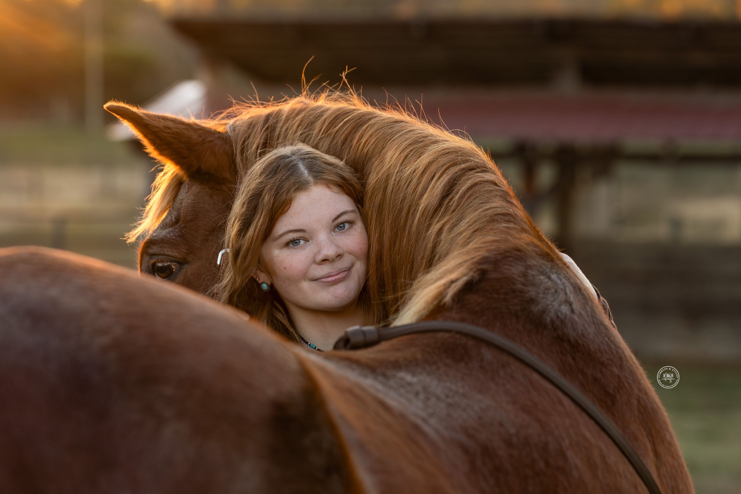 Young girl smiling while resting her head on a chestnut horse's neck at golden hour on a farm.