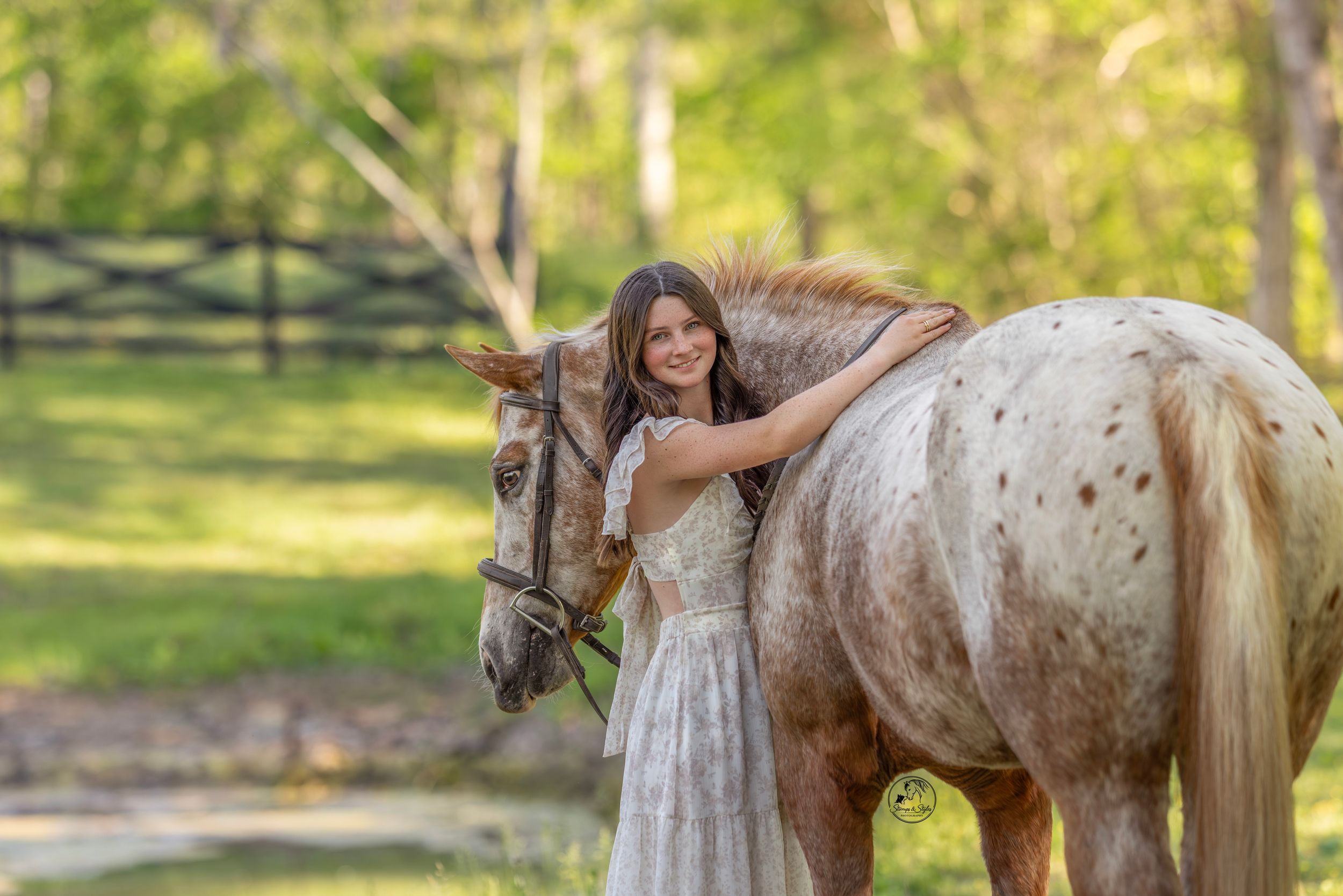 Young woman in white dress embracing a spotted Appaloosa horse outdoors in a lush green setting.