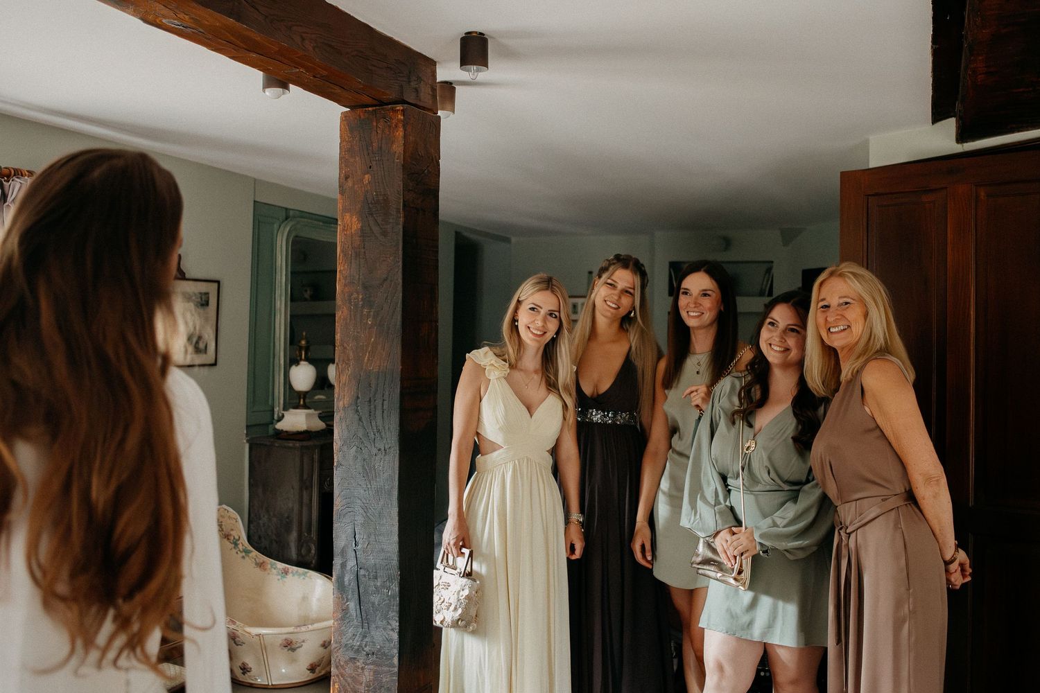 Bride in white gown smiles with bridesmaids in elegant dresses inside a rustic venue with wooden beams.