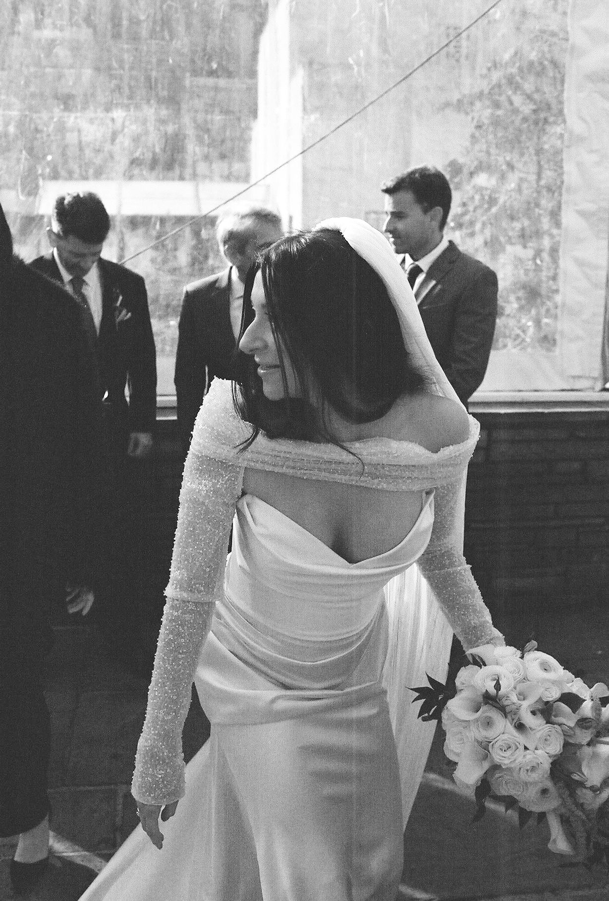 Bride and groom in wedding attire petting their black Labrador dog wearing a white floral collar indoors.
