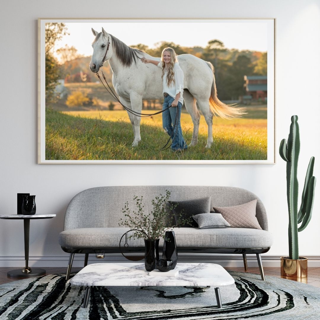 A woman in equestrian attire stands beside a chestnut horse in a sandy arena, displayed as framed wall art in a modern living room.