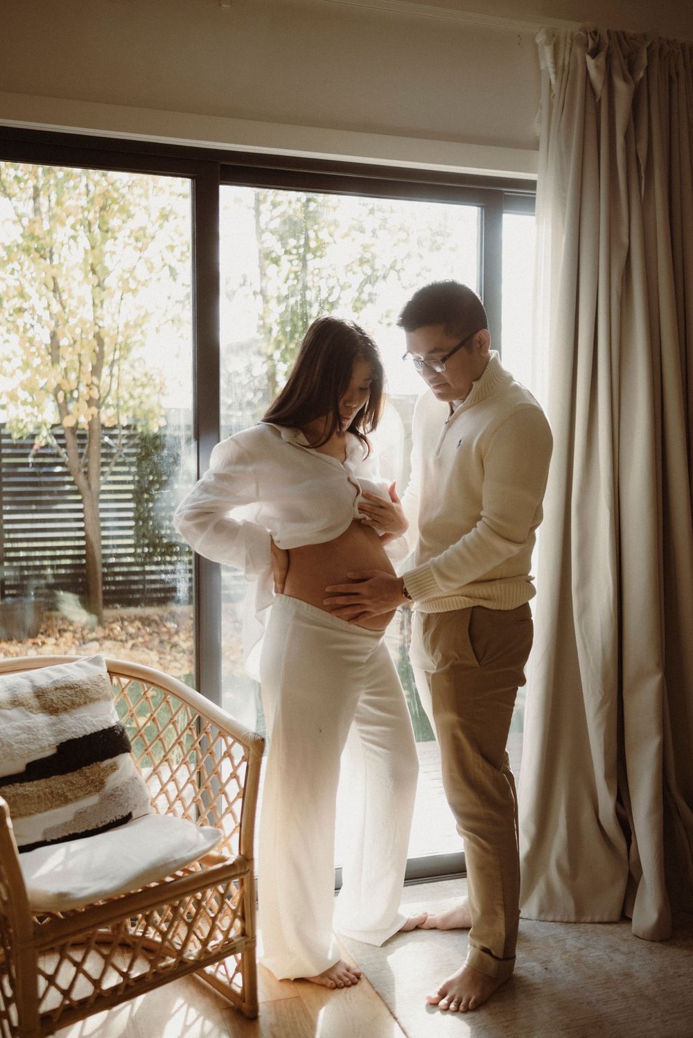A couple shares a tender moment as they cradle a pregnant belly together, standing by a sunlit window in a cozy room.