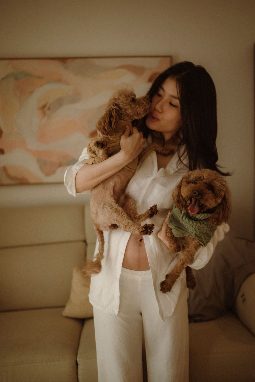 Pregnant woman in white outfit kissing and holding two brown poodle dogs at home.