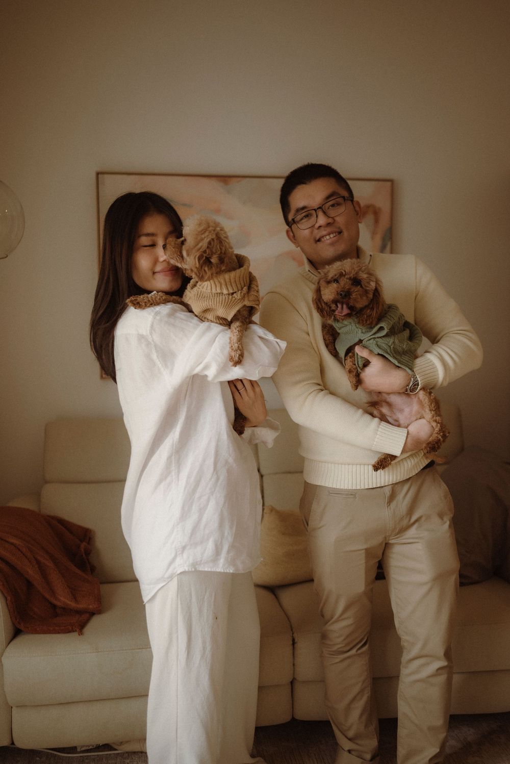 A couple holding two small dogs in sweaters, posing in a cozy living room with neutral tones.