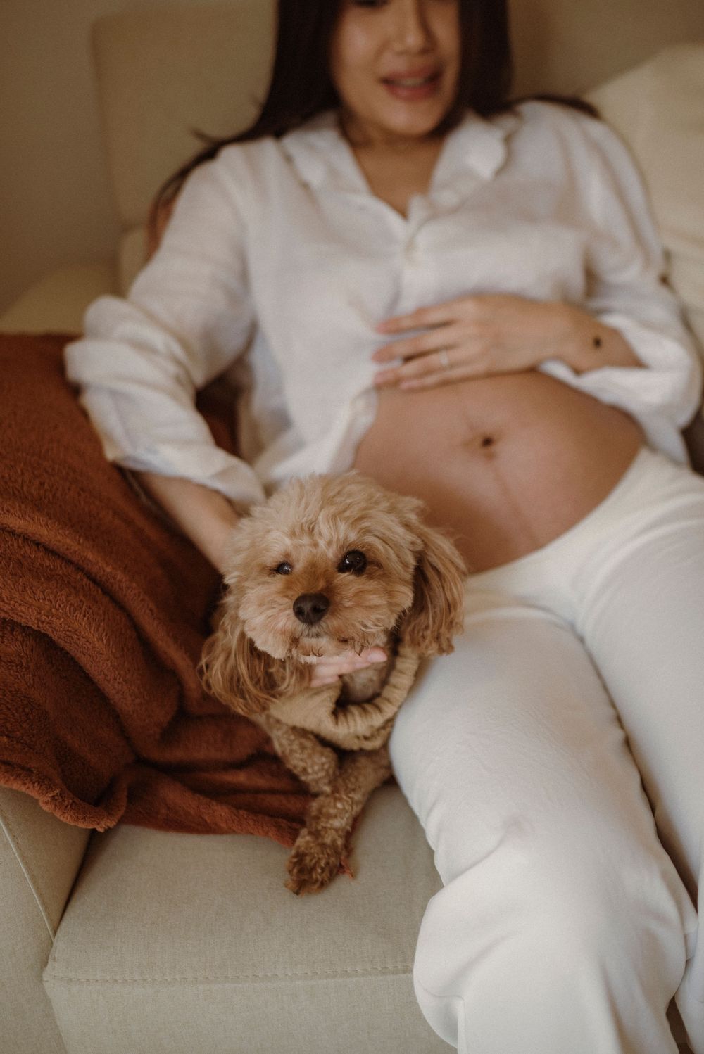 Pregnant woman in white outfit cradling baby bump while cuddly brown toy poodle sits beside her on a cozy sofa.