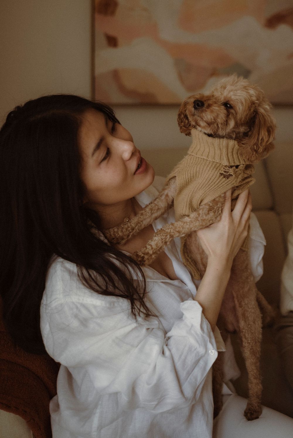 A young Asian woman in a white shirt holds a small brown poodle wearing a tan sweater indoors.